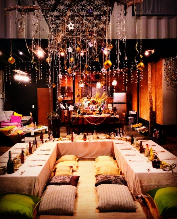 A spacious dining area is arranged with a large table set for a communal meal, adorned with decorative items and soft pillows for seating. Above, hanging ornaments and lights create a festive atmosphere. The kitchen area is visible in the background, enhancing the welcoming environment.