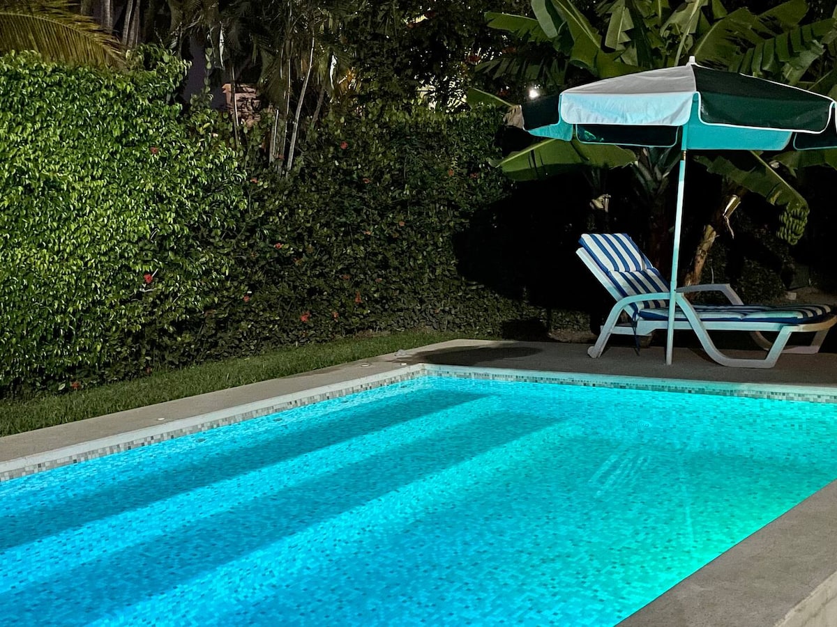 A private pool is illuminated in soft blue lights at night, surrounded by lush greenery. A shaded area with a striped umbrella and a lounge chair sits beside the water, creating a tranquil space for relaxation.
