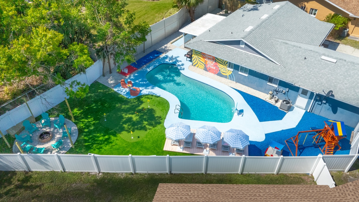 A spacious outdoor area features a sparkling swimming pool with a colorful deck. Surrounding the pool, seating areas include sun loungers and a fire pit. A playset is visible alongside the pool, and vibrant murals add a playful touch to the backyard.