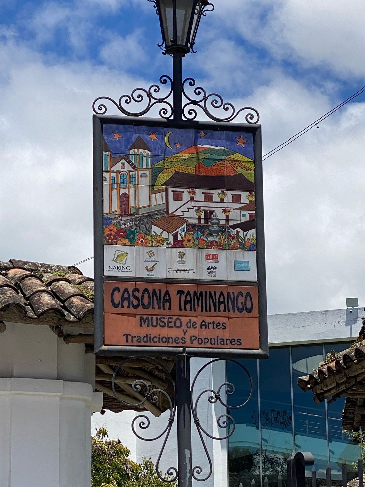 A colorful sign for Casona Taminango is displayed, showcasing artistic depictions of traditional scenes. The sign includes the name of the museum, 'MUSEO de Artes Tradiciones Populares,' and is framed by a picturesque street lamp and a building facade with clay tile roofing.