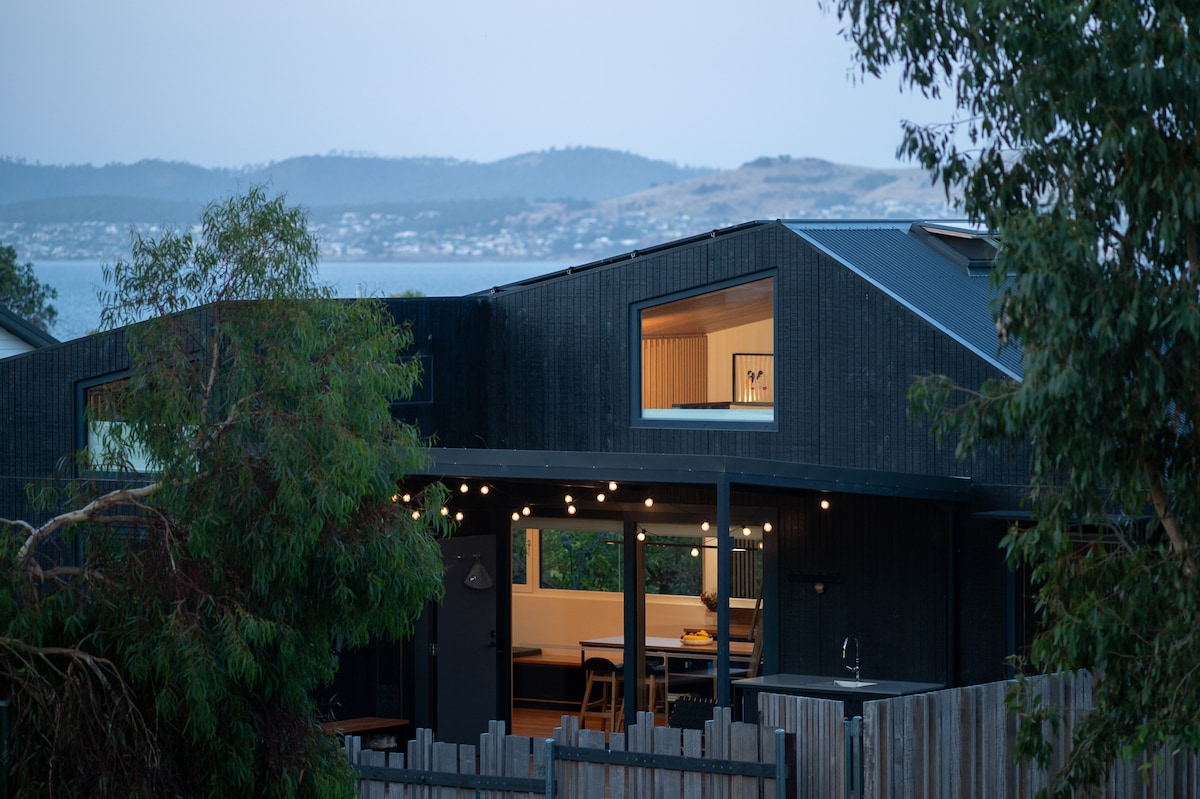 The exterior of a modern two-story home is shown, featuring a dark facade with large windows. Surrounding trees and a wooden fence enhance the natural setting, while distant hills and the River Derwent are visible in the background, creating a serene atmosphere.