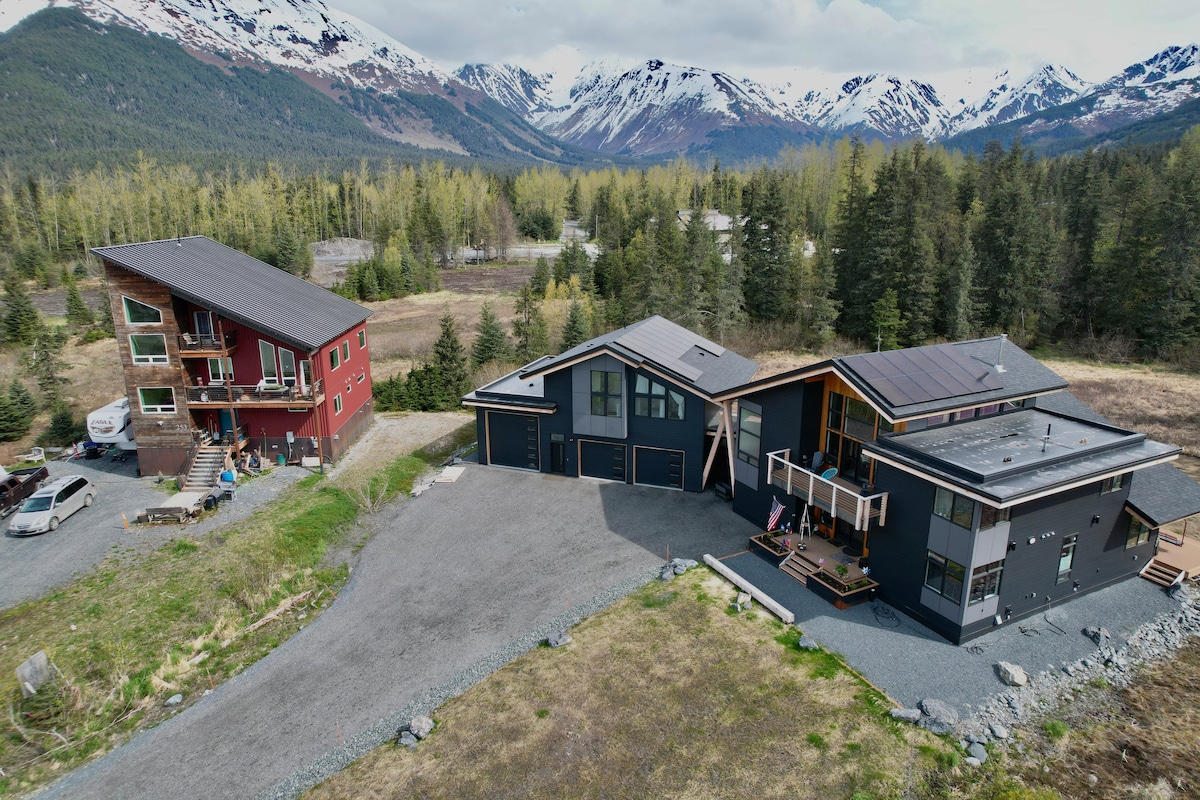 Aerial view of two modern buildings set against a backdrop of snow-covered mountains. The structures are surrounded by greenery and gravel pathways, showcasing a spacious outdoor area that invites exploration.