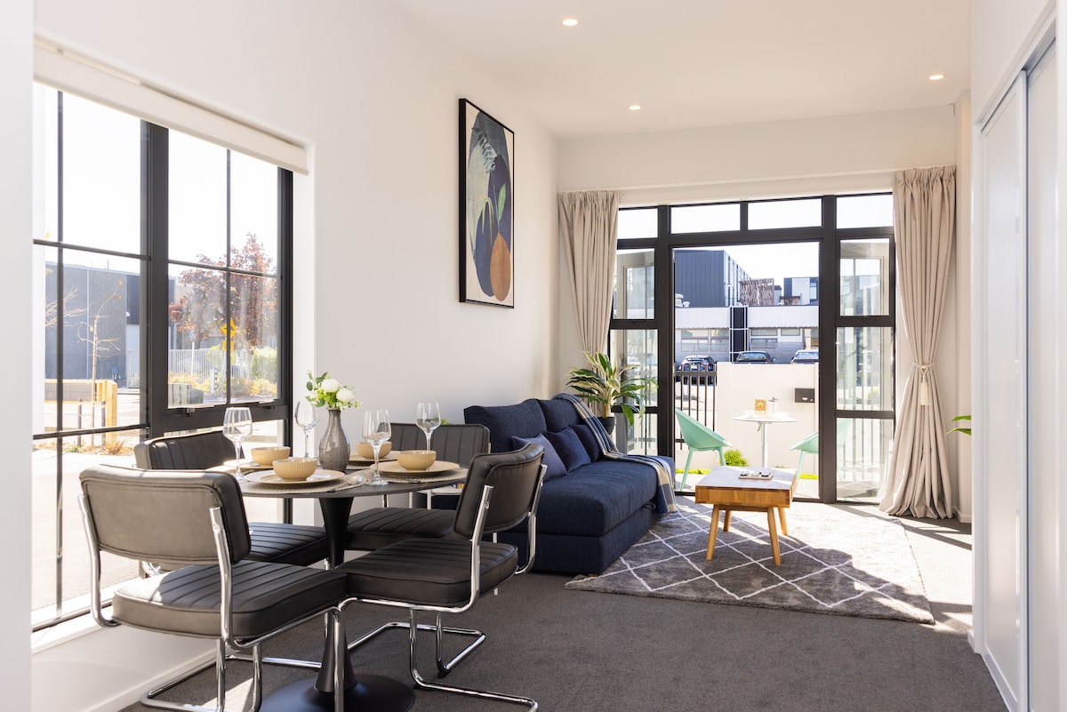 An open-plan living and dining area is illuminated by natural light, featuring a comfortable blue sofa and a round dining table with seating for four. Large windows provide views of the surroundings, while decorative plants enhance the space. A casual coffee table sits atop a textured area rug.