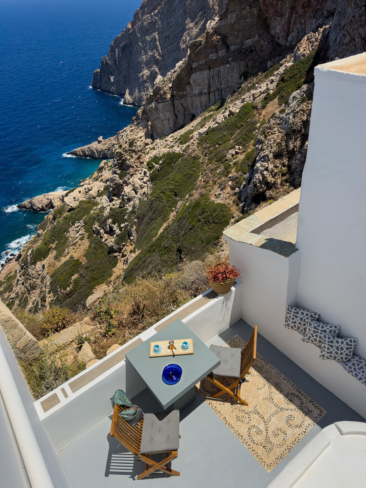An outdoor veranda with a small table set for two chairs overlooks a rocky coastline and the Aegean Sea. The table is decorated with a blue dish, and a potted plant adds a touch of greenery. The cliffside setting provides a sense of tranquility.