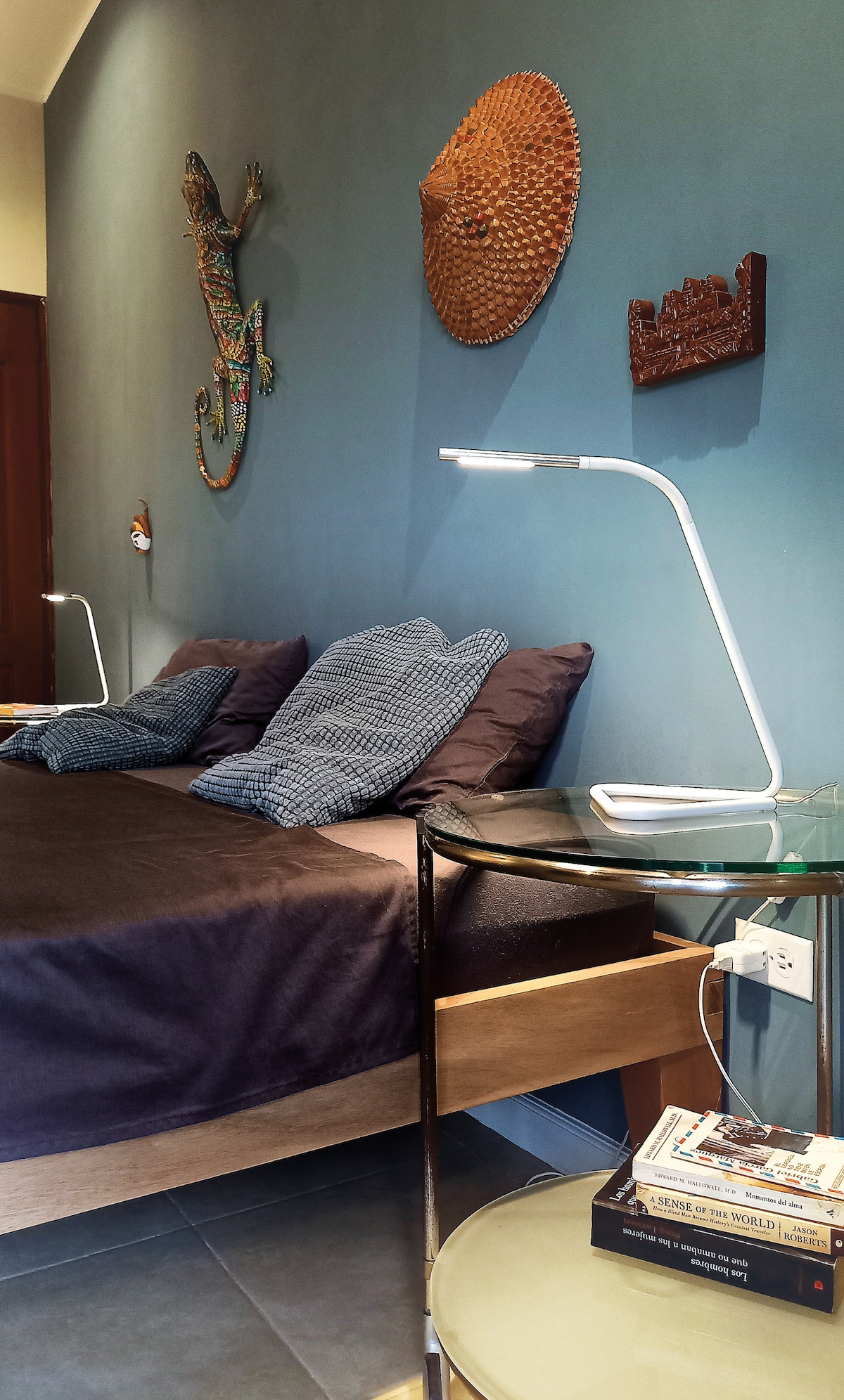 A spacious bed adorned with dark bedding and two patterned pillows is positioned against a teal wall. Decorative wall art, including a vibrant lizard and woven crafts, adds visual interest. A modern bedside lamp and a glass side table with books are also present.