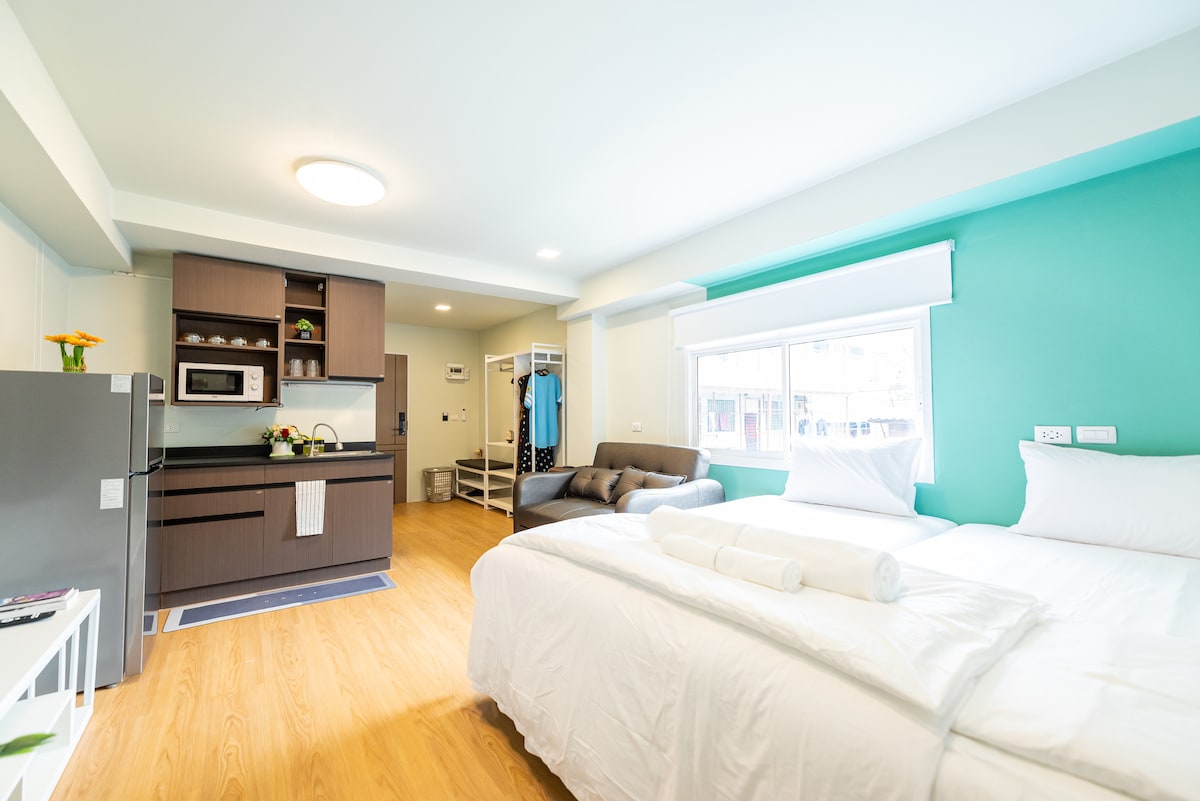 A bright and airy studio apartment features a spacious sleeping area with a neatly made bed and a cozy sofa. A fully equipped kitchenette is visible, complete with a microwave and dining area. Natural light filters in through large windows, complementing the soft blue walls.