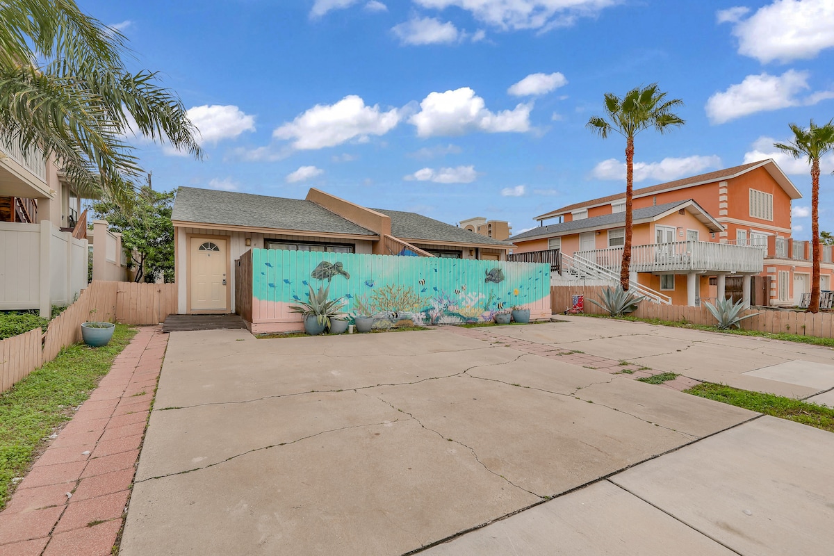 A large outdoor space is showcased with a vibrant mural featuring marine life on the front wall. Surrounding palm trees add a tropical feel, and a concrete driveway provides ample parking space for guests. The setting invites relaxation and connection to the coastal environment.