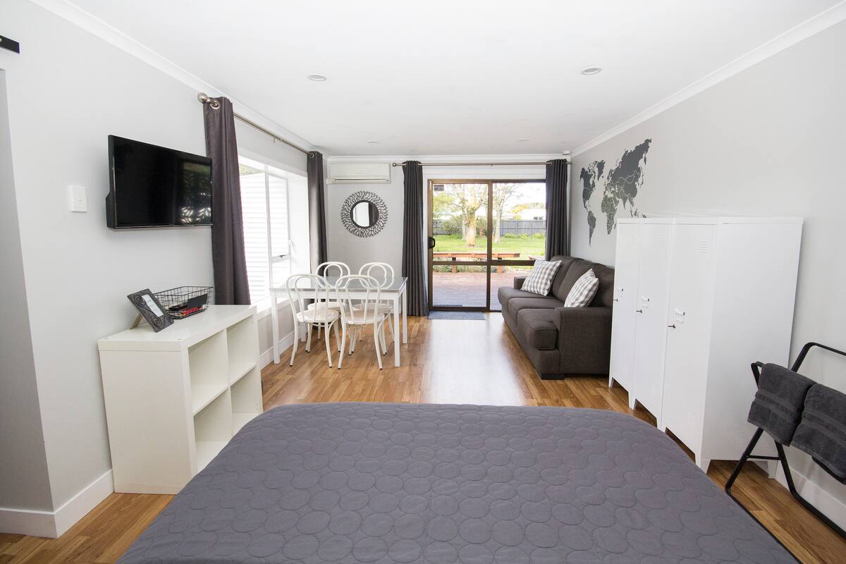 A modern studio space is shown with a comfortable bed and a cozy sofa. A dining table with chairs is positioned near large windows, allowing natural light. A TV is mounted on the wall, while a sleek white cabinet provides storage.