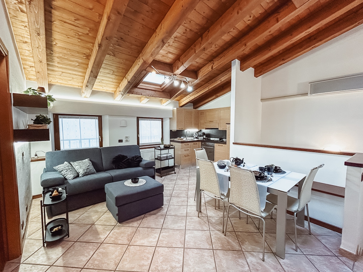 The open-plan living area features high wooden beams overhead and a cozy lounge space with a dark sofa and ottoman. A dining table with chairs is set in the foreground, accompanied by modern kitchen cabinetry and countertops showcasing a well-equipped cooking space.