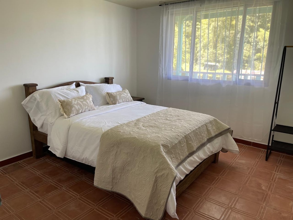 A spacious sleeping area features a wooden bed with a light-colored bedding set. Two decorative pillows are placed on the bed, and natural light streams in through a large window, partially covered by sheer curtains. The floor is finished with terracotta tiles.