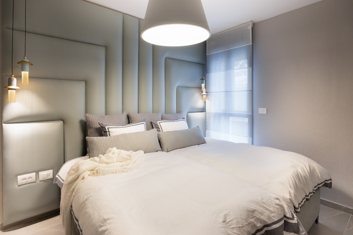 The bedroom features a spacious arrangement of two beds with soft bedding, highlighted by subtle textures on the wall. Natural light filters through sheer curtains, while modern pendant lights add elegance and a warm glow to the overall ambiance.