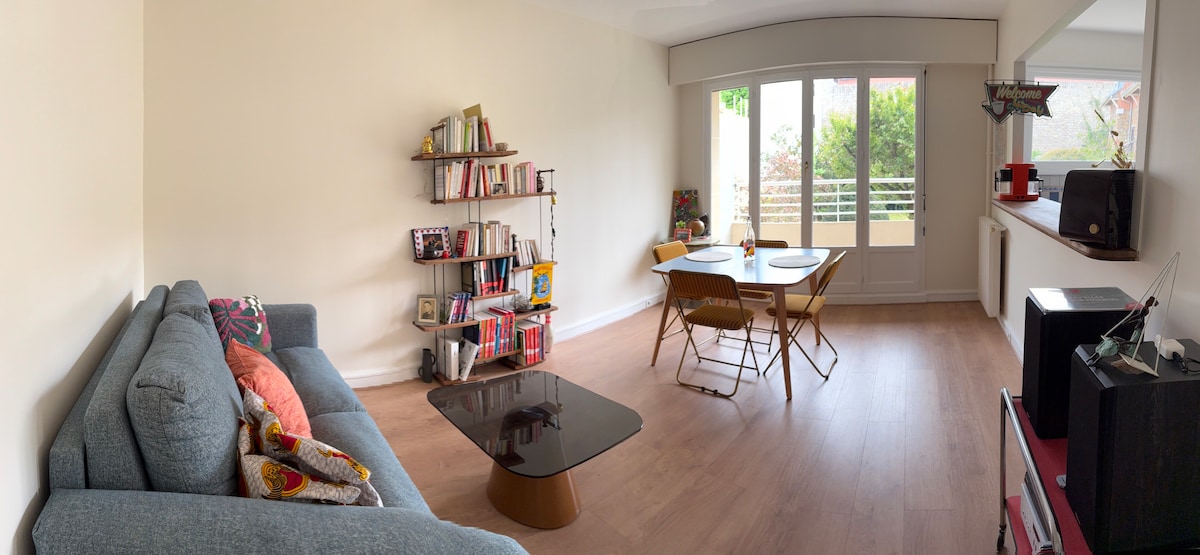 A light-filled living and dining area features a comfortable sofa, a low coffee table, and a small dining set. A bookshelf filled with various books adds character to the room. Large windows provide garden views, enhancing the bright and open feel of the space.