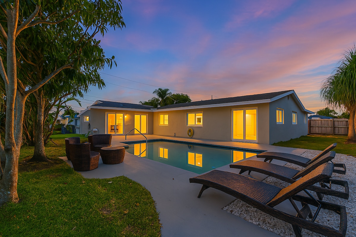An outdoor area features a private pool reflecting the evening sky, framed by lush greenery. Sun loungers are arranged along the pool's edge, while a seating arrangement is situated nearby, creating a welcoming space for relaxation. Warm lights glow from the windows of the home.