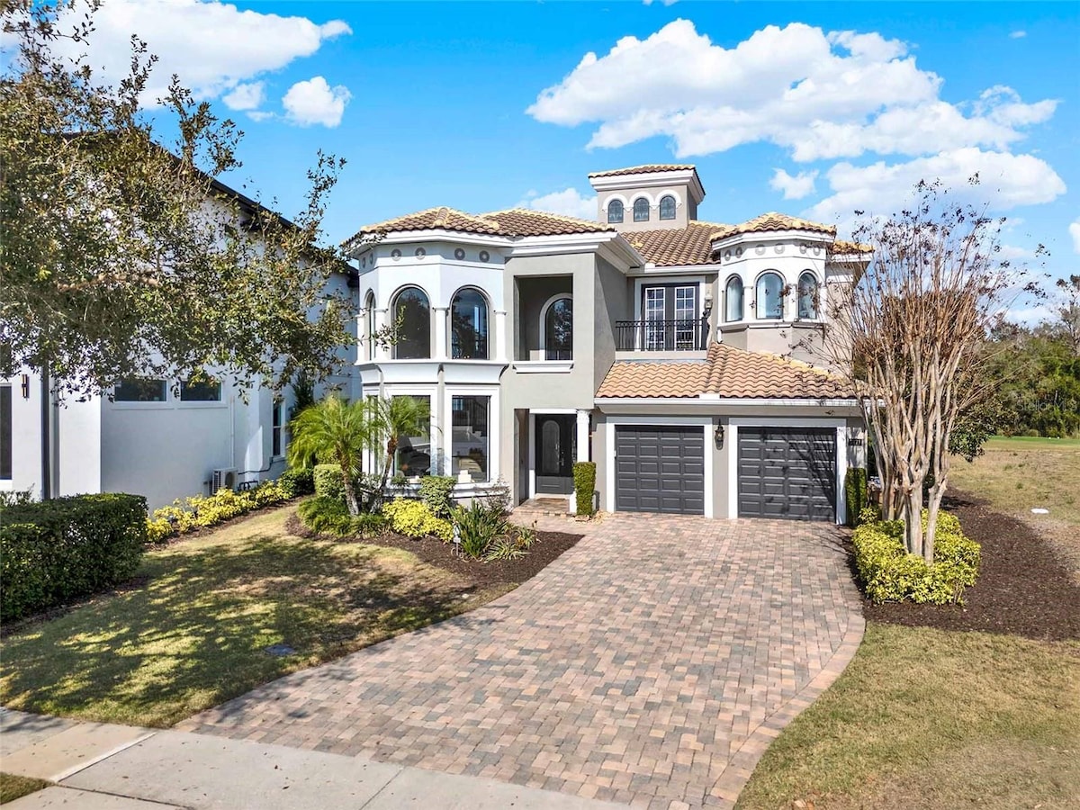 A luxurious vacation home is showcased from the front, featuring an elegant facade with arched windows and a tile roof. The driveway is paved with bricks, flanked by well-maintained landscaping that includes shrubs and palm trees.