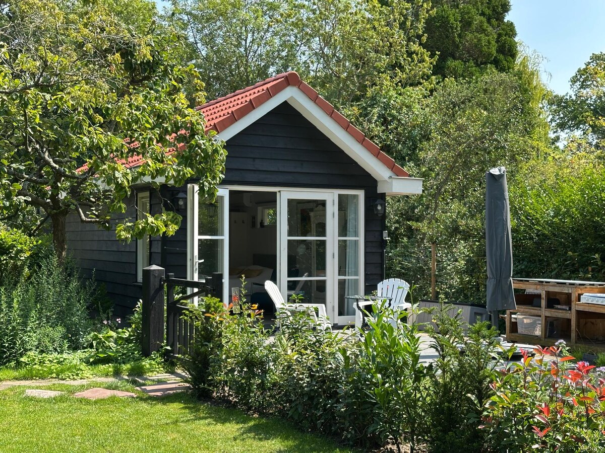 A quaint wooden cabin is framed by lush greenery and colorful flowers. Large windows invite natural light and provide views of the outdoor patio, equipped with several chairs and a sun umbrella. The red-tiled roof contrasts with the dark wood of the structure.