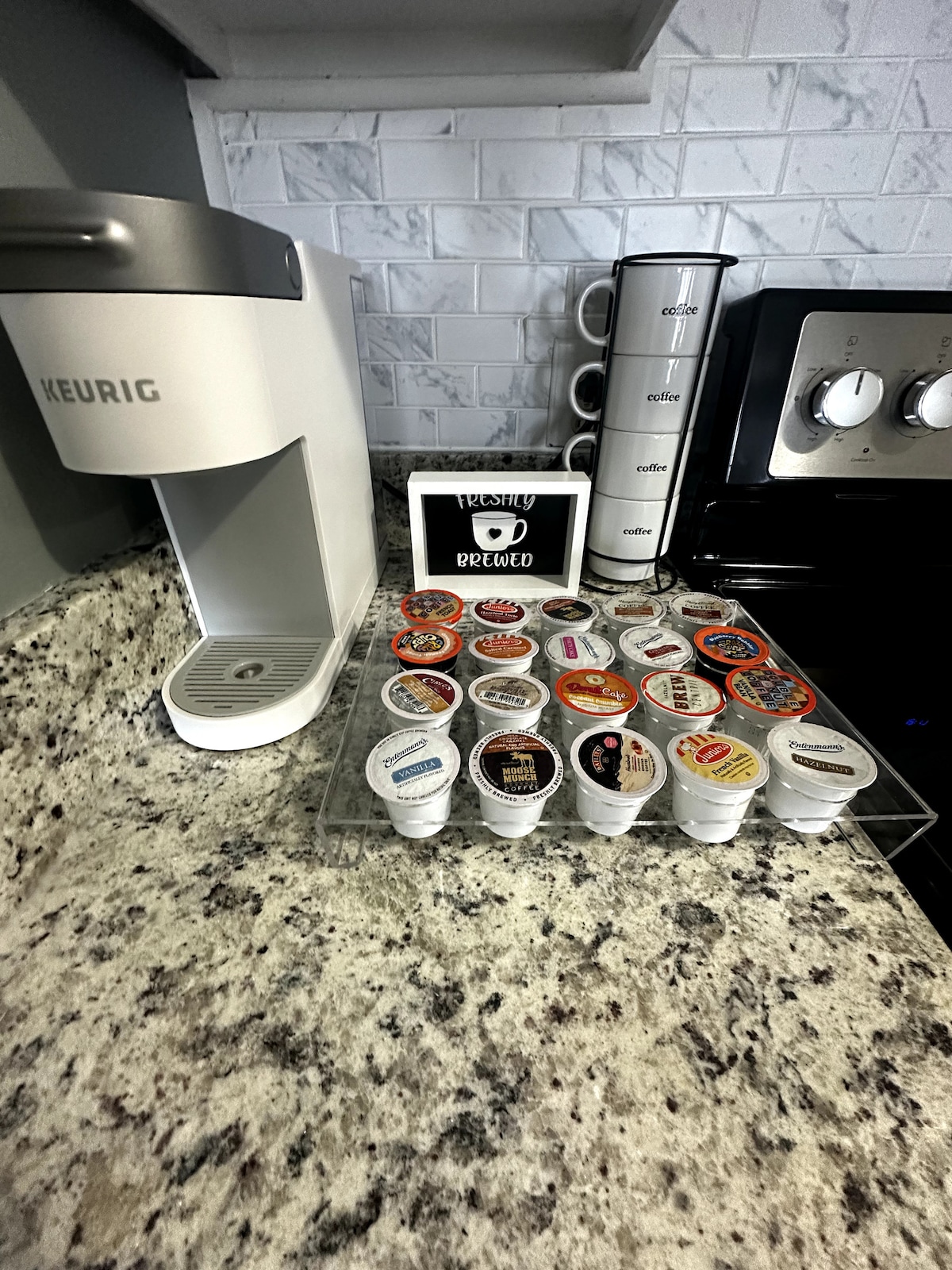 A coffee station is displayed on a granite countertop, featuring a Keurig coffee maker alongside a variety of coffee pods. The pods are organized in clear containers, with a small sign that reads 'Coffee Brewed' positioned nearby.