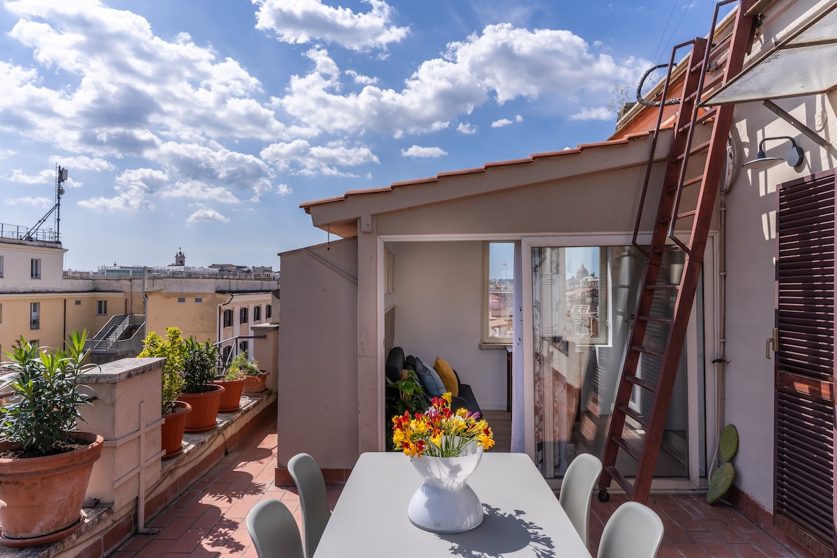 A private terrace is presented, featuring a dining table surrounded by chairs and a vibrant flower bouquet in the center. Lush plants adorn the edges, and a wooden ladder leads to the rooftop, with a backdrop of a bright blue sky and scattered clouds.