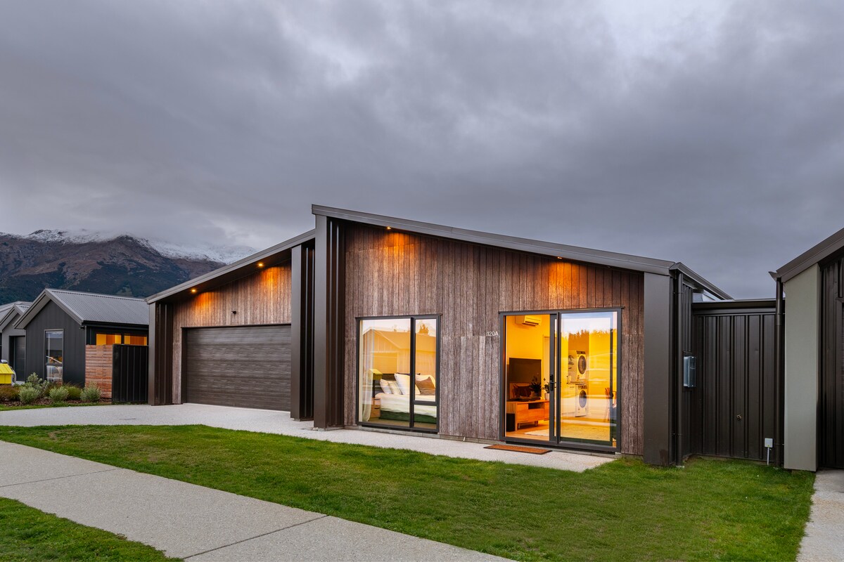 The exterior of the Lake Hāwea Luxe Suite showcases a modern design with expansive windows, offering views of the interior. The charming facade features natural wood accents and soft lighting, while the neatly landscaped surroundings highlight a welcoming approach to the private entrance.