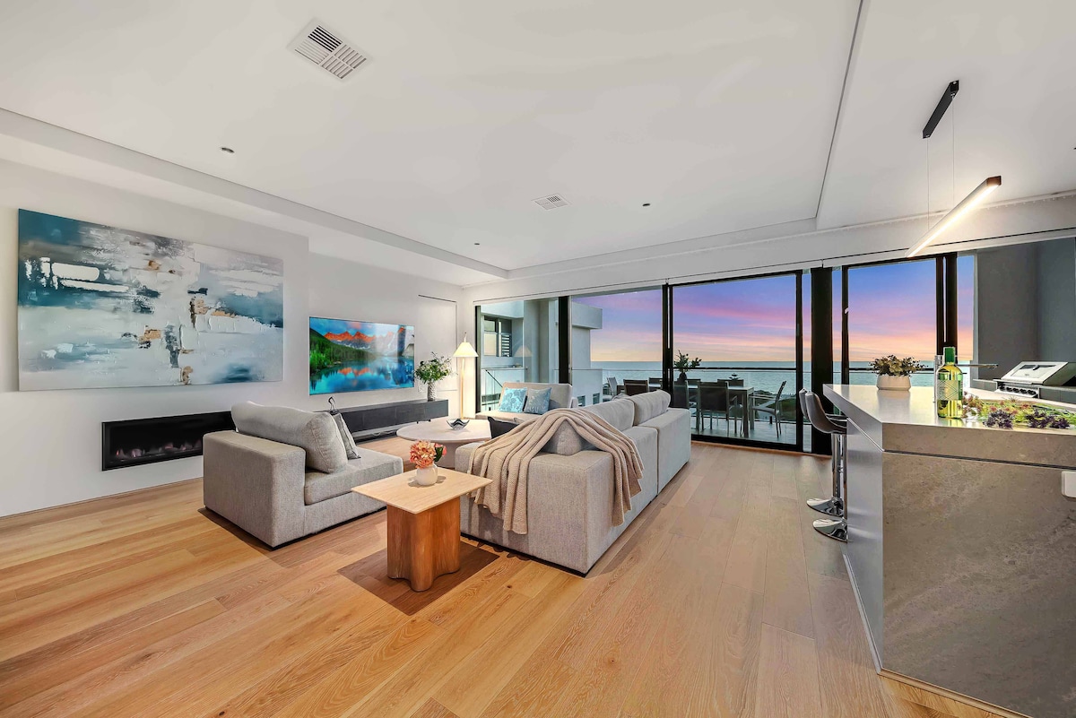 An open-plan living area features modern furnishings with a cozy sofa and stylish coffee table. Large glass doors provide views of the ocean, allowing natural light to fill the space. Artwork adorns the walls, complementing the contemporary design and serene atmosphere.