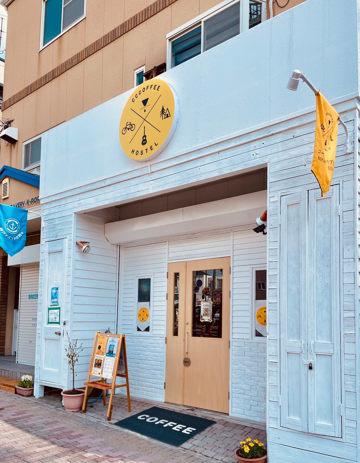 The exterior of CCCOFFEE HOSTEL features a light blue façade with white trim. A large circular sign displays the hostel's name and logo. Two potted plants are placed near the entrance, which is framed by wooden doors. Colorful flags are visible on either side.