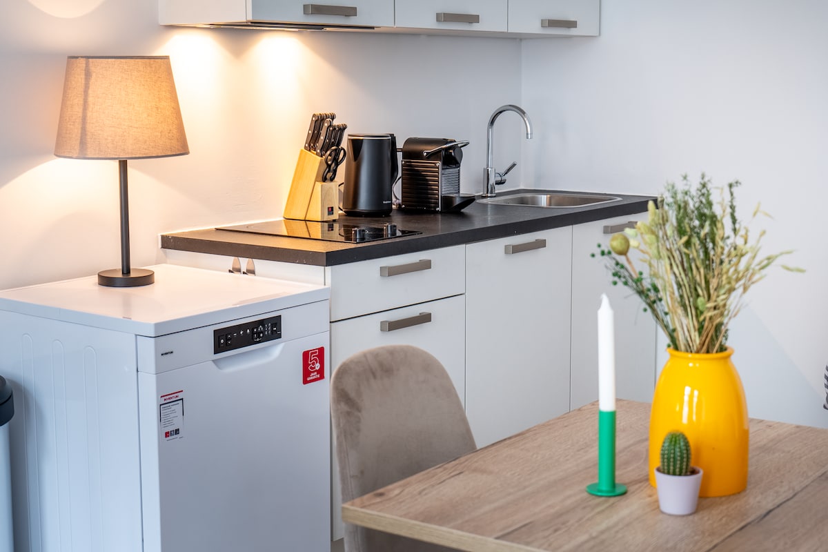 A modern kitchen area is featured, equipped with essential appliances including a refrigerator and coffee maker. A well-lit countertop displays cooking utensils and a lamp. The dining table, set with a vibrant vase and a candle, offers a cozy space for meals or work.