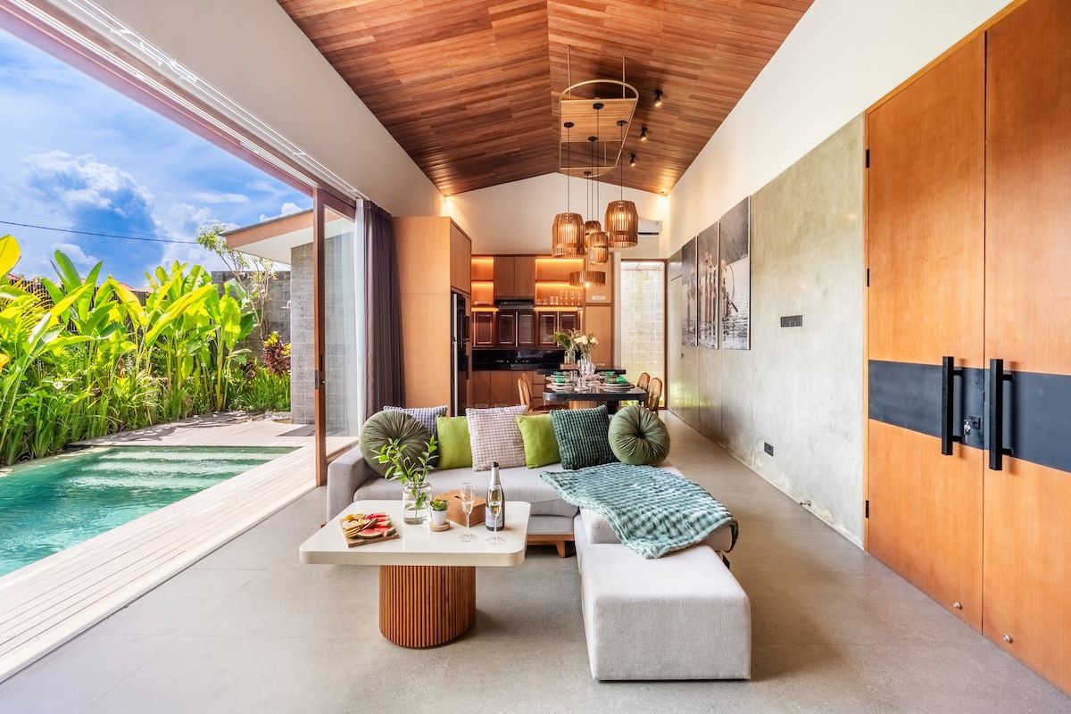 An open living space features a modern design with a high wooden ceiling. A comfortable sofa is arranged around a light wood coffee table, accompanied by decorative items. Floor-to-ceiling glass doors provide a view of the vibrant garden and private pool, inviting natural light indoors.