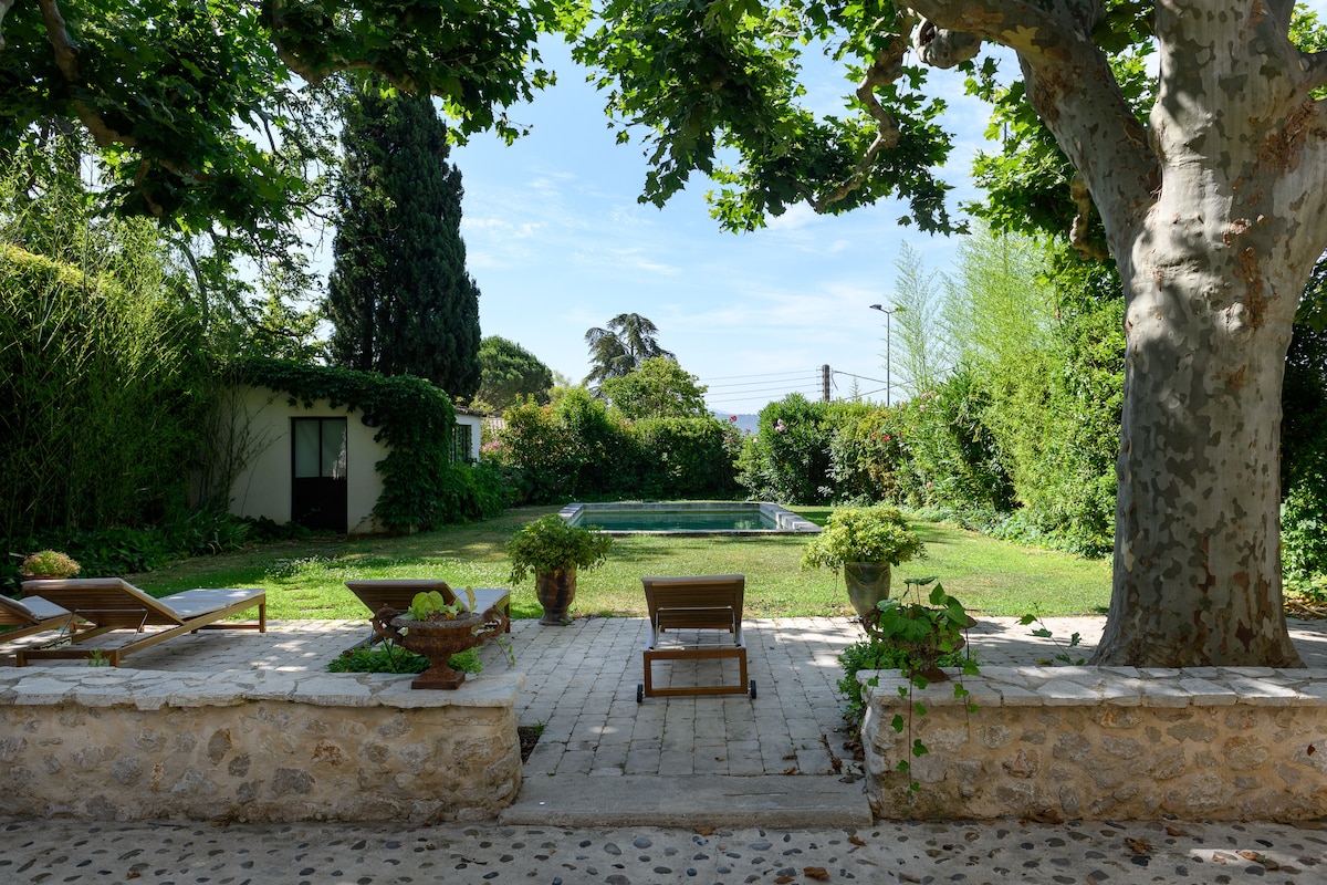 A serene garden space is seen featuring lush greenery and mature trees. Lounge chairs are positioned on stone paving, providing a view of the inviting swimming pool. The area exudes a sense of tranquility, with sunlight filtering through the foliage.