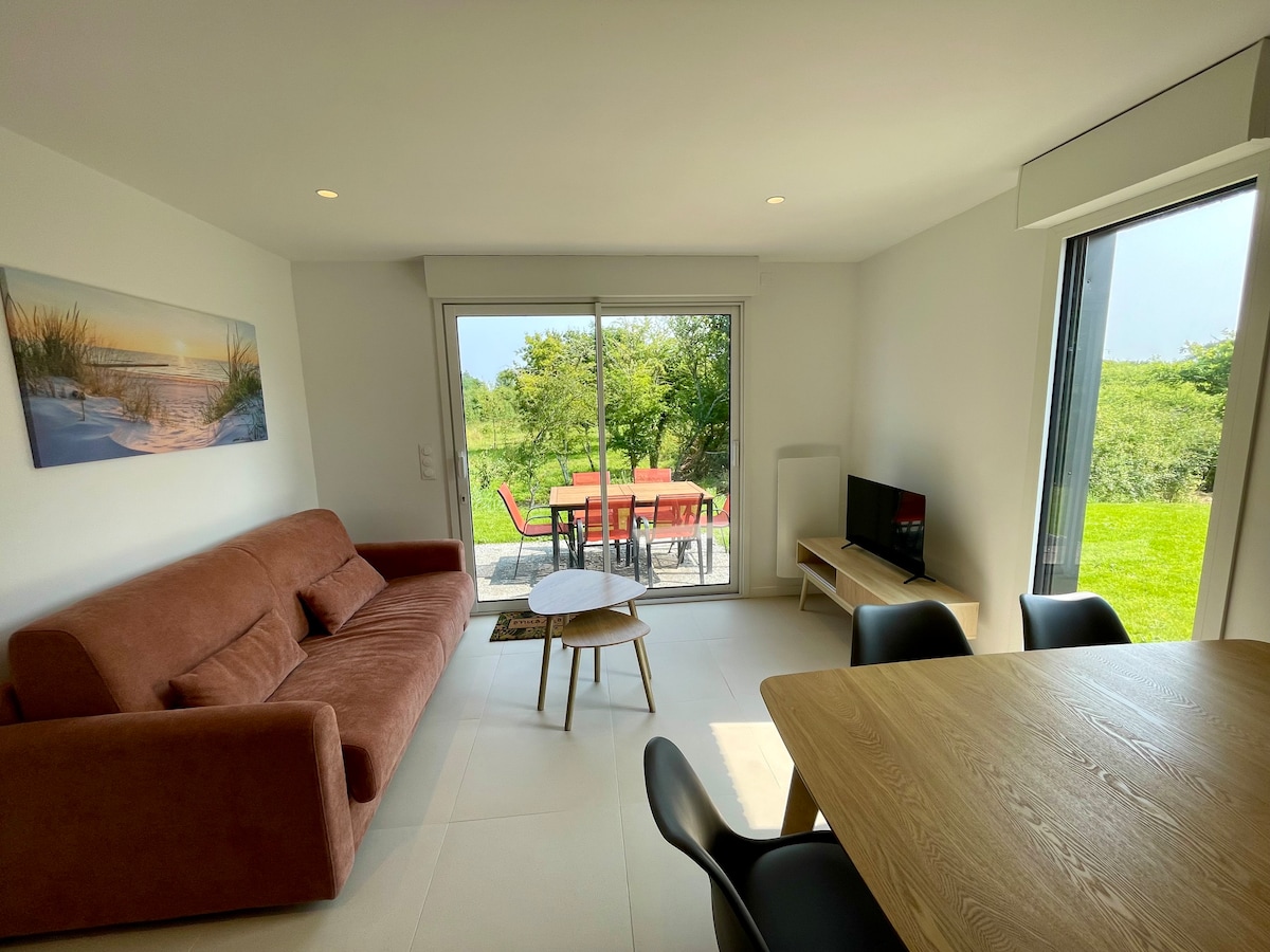 A living area features a convertible sofa and a small wooden coffee table. Large glass doors provide a view of the outdoor terrace and garden area. A dining table with black chairs is positioned against the wall, alongside a television set.
