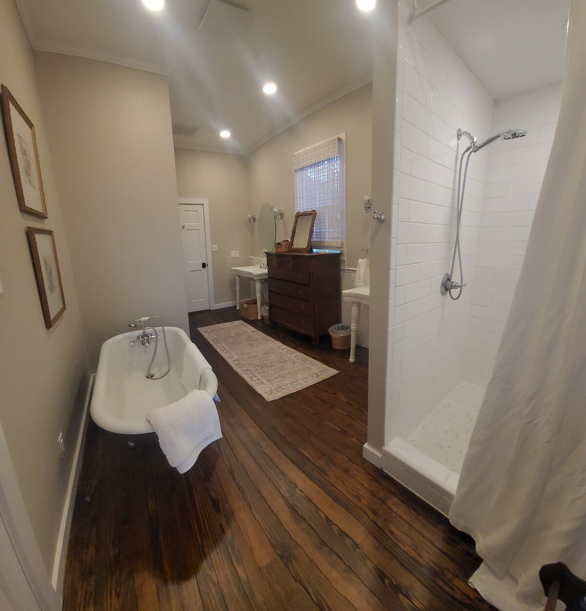 A well-lit bathroom features a classic freestanding bathtub alongside a walk-in shower. Natural wood flooring adds warmth, while a simple area rug enhances the space. A large mirror and bathroom accessories provide functionality, contributing to the overall inviting ambiance.