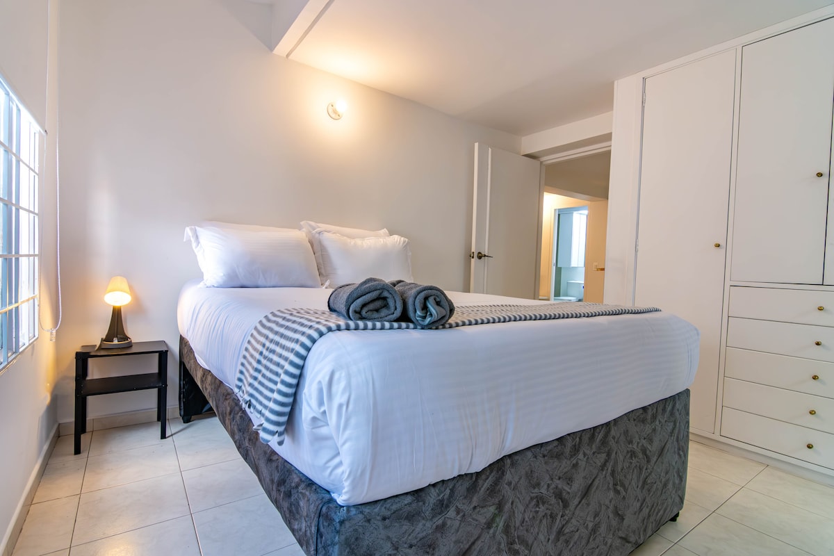 A well-appointed bedroom features a spacious bed topped with crisp white linens and neatly arranged gray towels. A bedside table with a soft lamp adds a warm touch. Bright natural light filters through a nearby window, while a closet and door to the bathroom are visible.