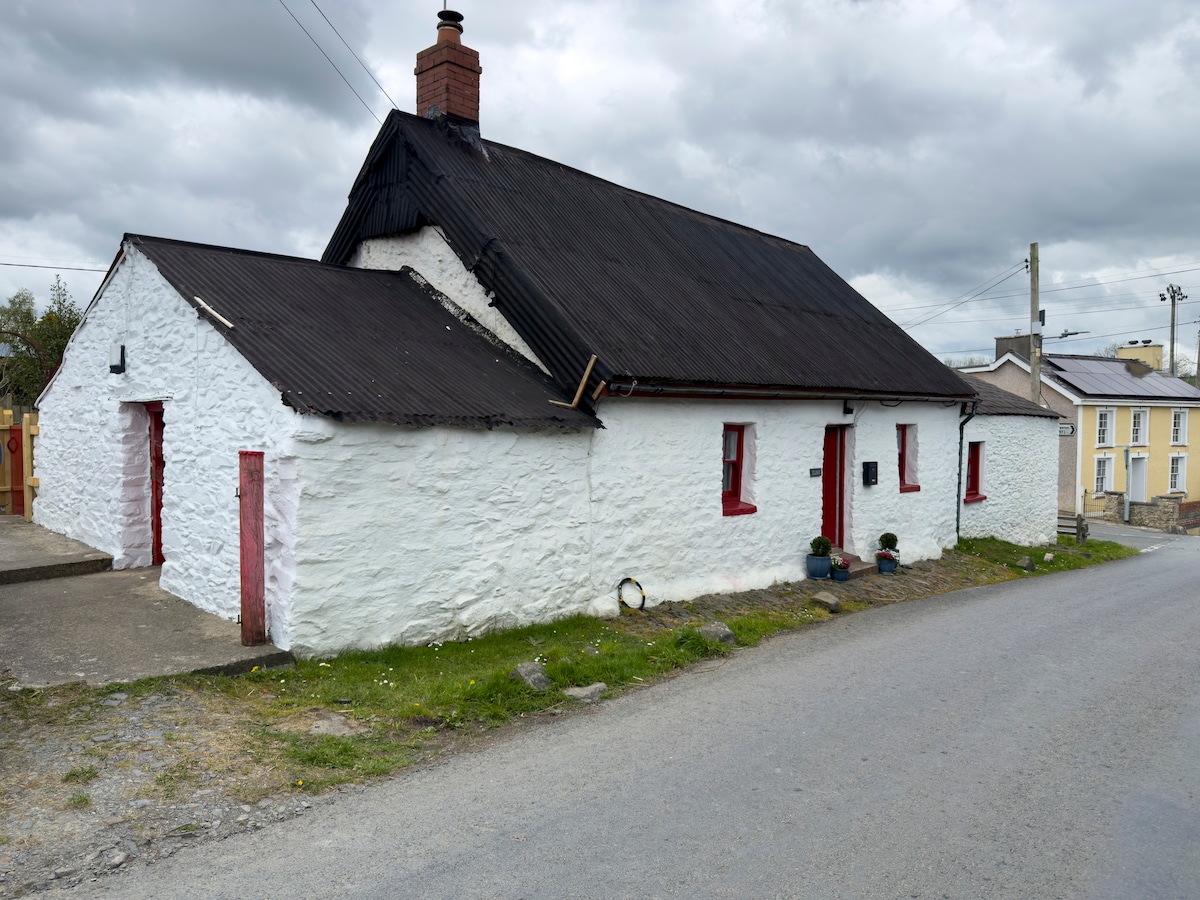 A charming white cottage features a distinctive black roof and red door accents. Positioned along a quiet street, the weathered stone exterior highlights its rustic character. Surrounding greenery contributes to the rural setting, offering a quaint and inviting appearance.