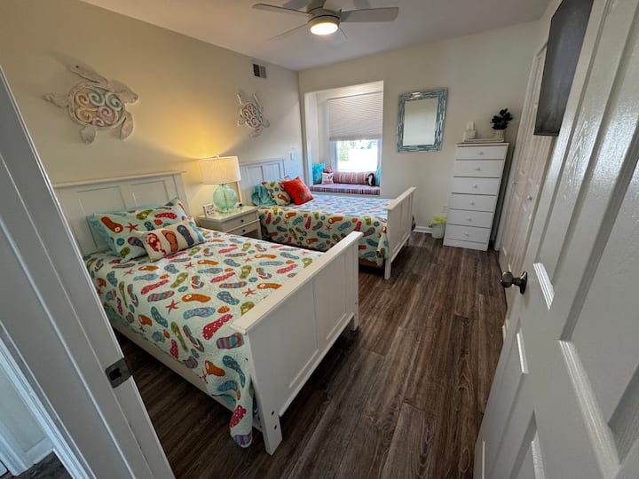 Ნათელი ტყუპი საძინებელი ფერადი პლაჟის თემატიკის თეთრეულით,  თეთრი ხის ავეჯითა და კუს კედლის ხელოვნებით. Აქვს ფანჯრის მყუდრო სავარძელი აქცენტიანი ბალიშებით, ფირუზისფერი ლამპა ტუმბოზე და ჭერის ვენტილატორი განათებით.