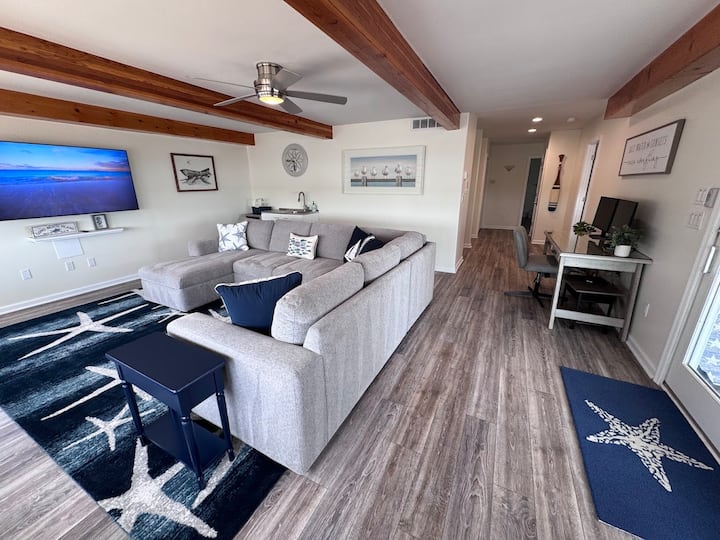 Ელეგანტური სანაპირო თემატიკის 1-ლი სართულის მისაღები ოთახი აღჭურვილია ფართო ნაცრისფერი სექციით, 65"-იანი სმარტ-ტელევიზორითა და ხის ჭერის დაუცველი კოჭებით.  Შესანიშნავი გასართობი სივრცე ბავშვებისთვის ან საოჯახო ფილმების ღამე.