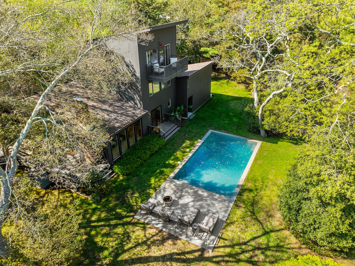 An aerial view showcases a modern retreat nestled within lush greenery. A rectangular outdoor pool is centered in the yard, surrounded by a stone patio that features lounge chairs. The home's exterior, characterized by large glass doors, complements the natural surroundings.