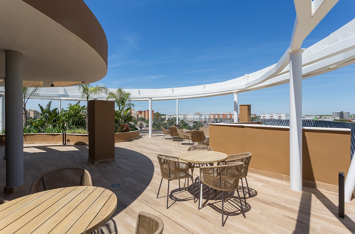 A spacious communal terrace is shown, featuring several round wooden tables surrounded by woven chairs. Lush greenery is present in planters, and an unobstructed view of the blue sky creates a bright and open atmosphere.