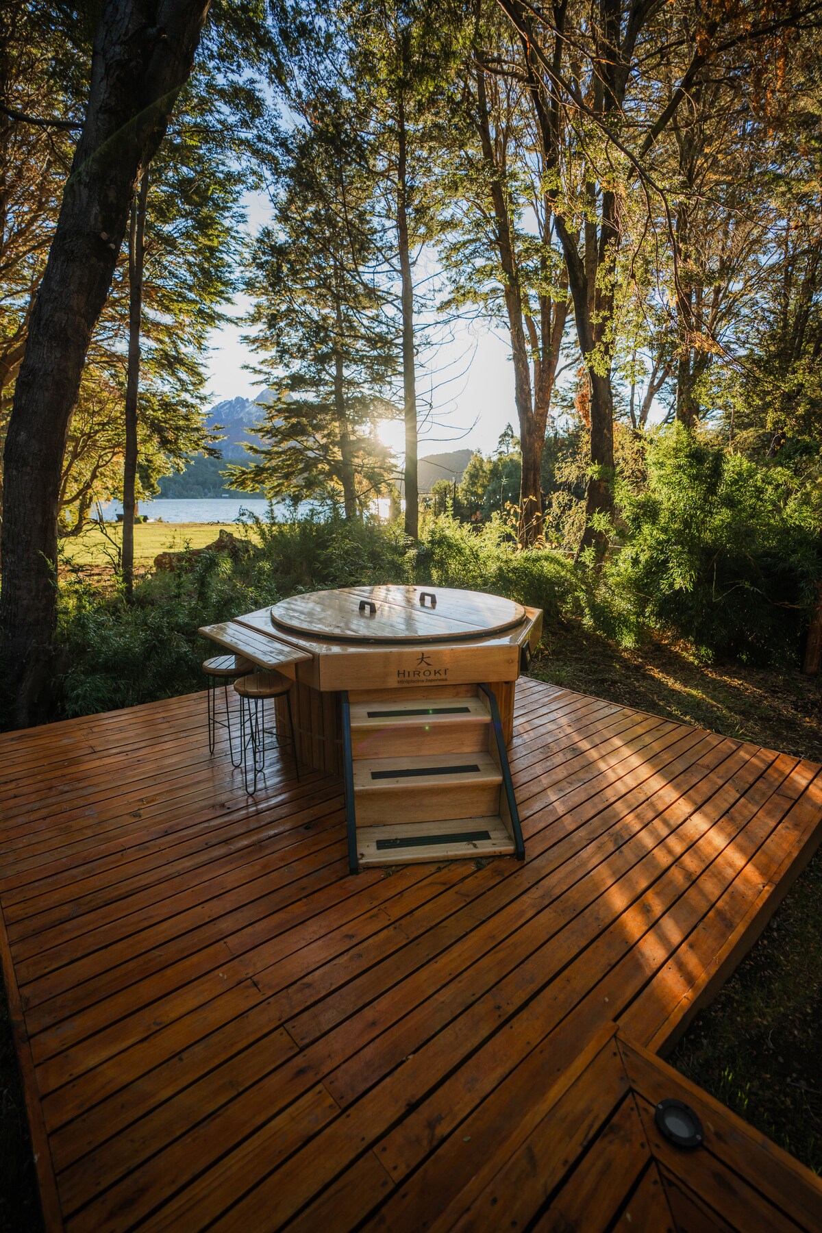A private wooden deck features a Japanese Hiroki bathtub surrounded by native trees. Sunlight filters through the branches, casting warm tones on the deck. The view of the lake adds a serene backdrop to this outdoor space, enhancing the connection with nature.