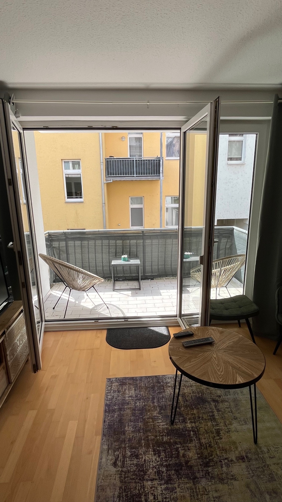 A balcony is visible through open double doors, revealing two outdoor chairs positioned on a tiled patio. A small round table with a decorative surface stands nearby, while natural light illuminates the interior space featuring wooden flooring and soft green accents.