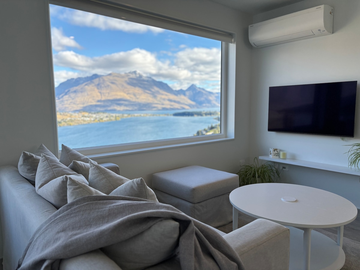 A modern living area features a large window framing breathtaking views of Lake Wakatipu and the surrounding mountain ranges. A cozy grey sofa with cushions and a matching ottoman sits adjacent to a small white coffee table, inviting relaxation while enjoying the scenery.