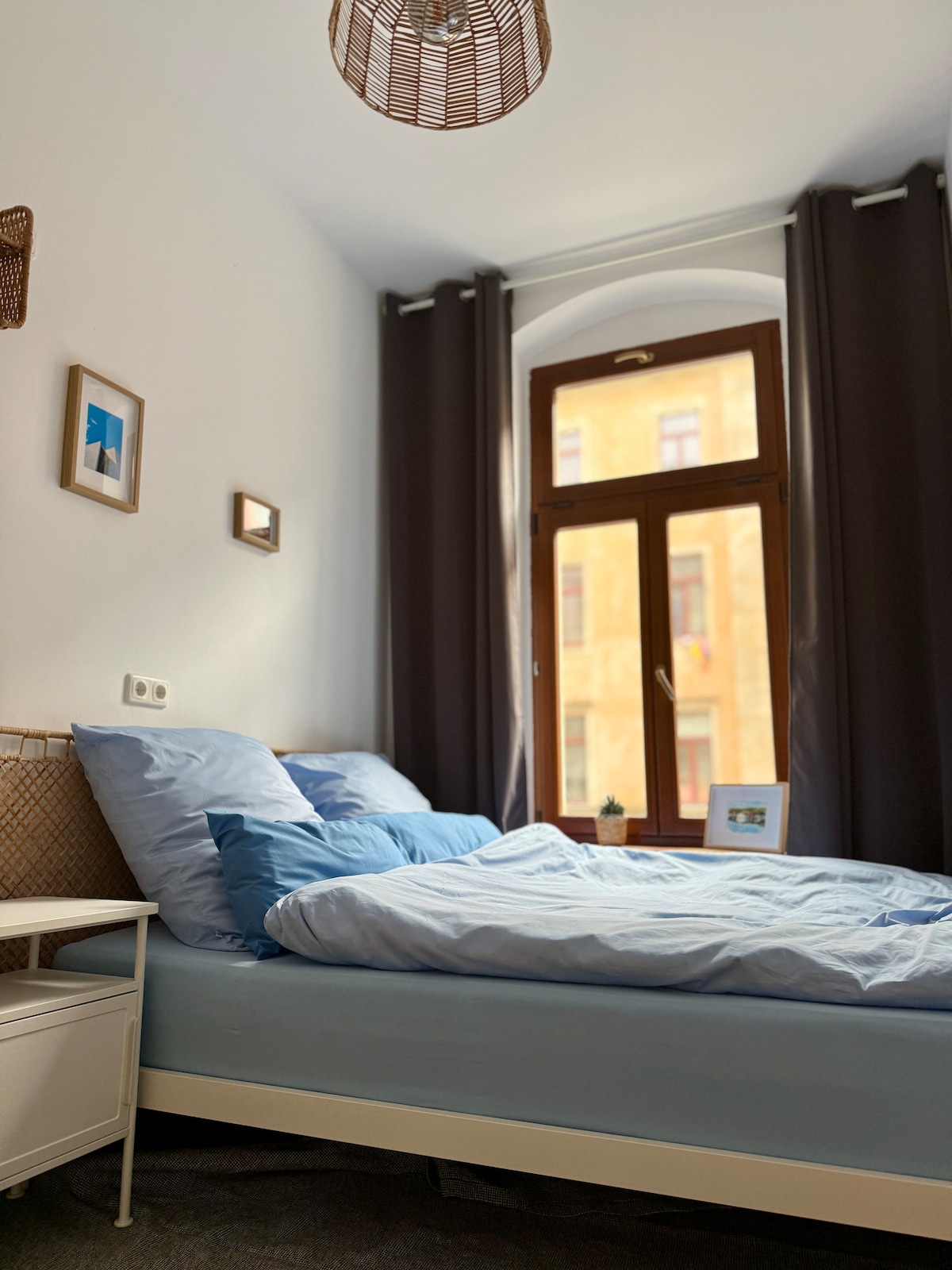 A cozy bedroom is presented with a high-quality queen-size bed dressed in soft blue linens. Natural light filters through a large window with darkening curtains. Simple decor, including framed images and a small potted plant, enhances the space's character.