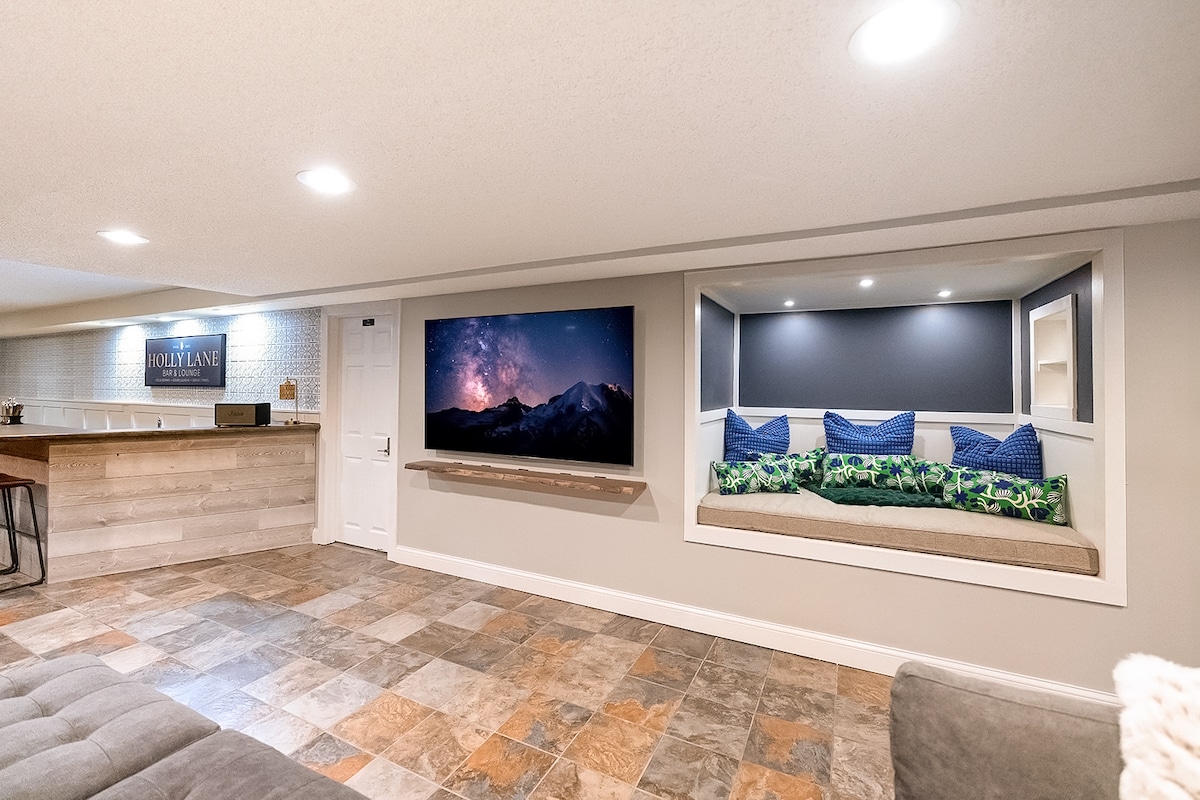 A spacious basement area is highlighted by a large television mounted on the wall, showing a scenic mountain landscape. A cozy nook with cushioned seating is visible, adorned with blue and green pillows. The bar area is in the background, featuring a wooden countertop.