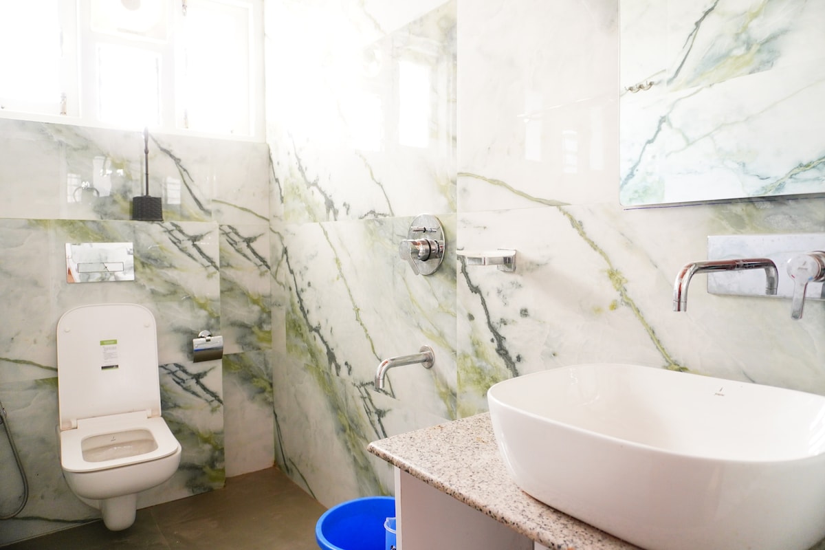 A modern bathroom features marble walls with green and white veining. A sleek, round washbasin sits atop a granite counter, while a contemporary toilet is placed to the side. A blue bucket is visible on the floor, and ample natural light fills the space.
