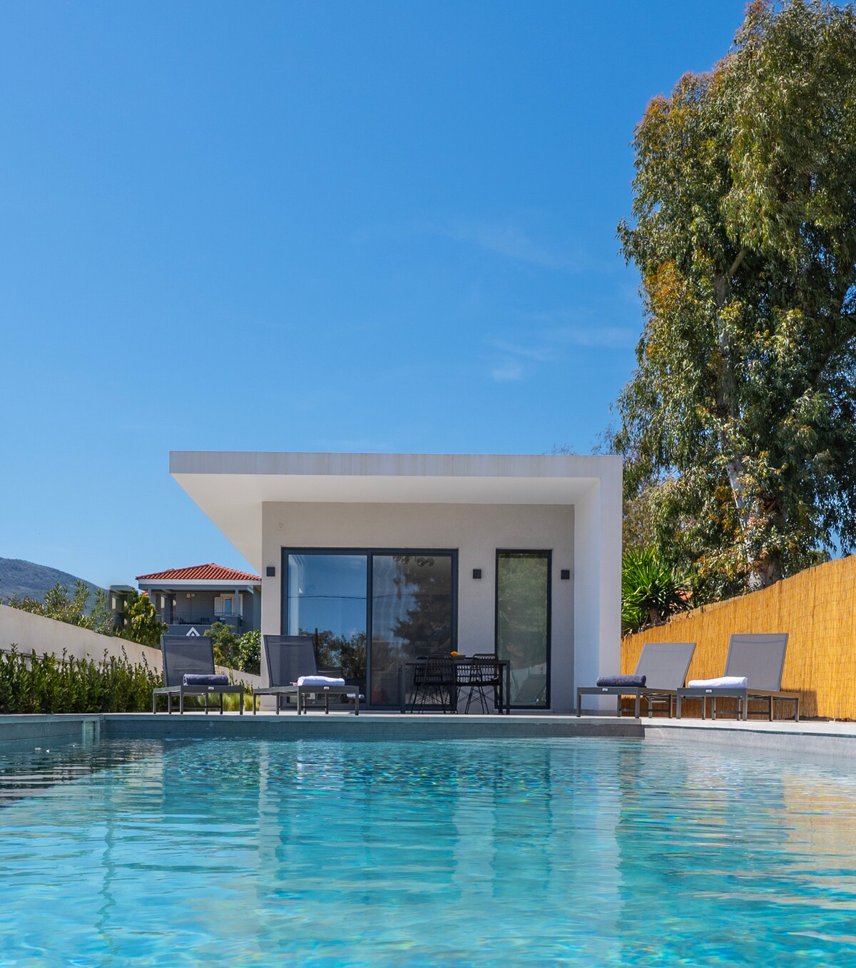 An inviting villa is framed by lush greenery and clear blue skies. A private pool reflects the sunlight and is surrounded by several lounge chairs. The sleek structure features large glass doors that open onto the outdoor space, enhancing the property's connection with nature.