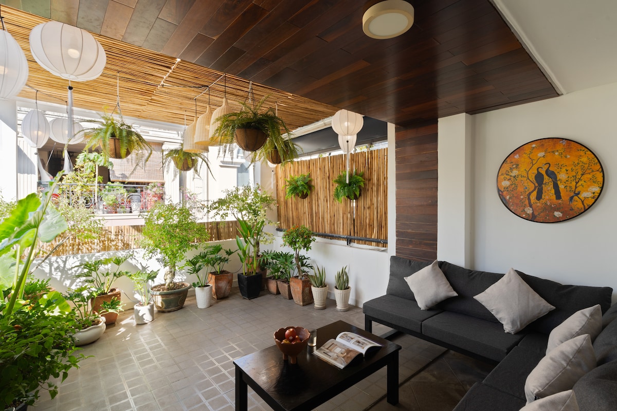 A tranquil outdoor space is revealed, featuring a dark sofa paired with a small coffee table. Lush green plants are displayed in various pots, creating a vibrant environment. Soft light is filtered through pendant lamps, enhancing the peaceful atmosphere of this inviting area.