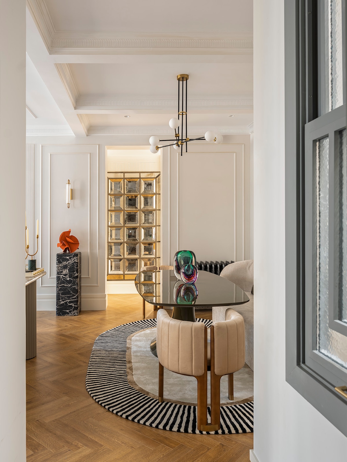 A modern dining area features a glass table surrounded by plush seating. A striking sculpture adds a pop of color, while the rug with bold patterns complements the space. Light wood flooring and intricate wall design enhance the sophisticated atmosphere.