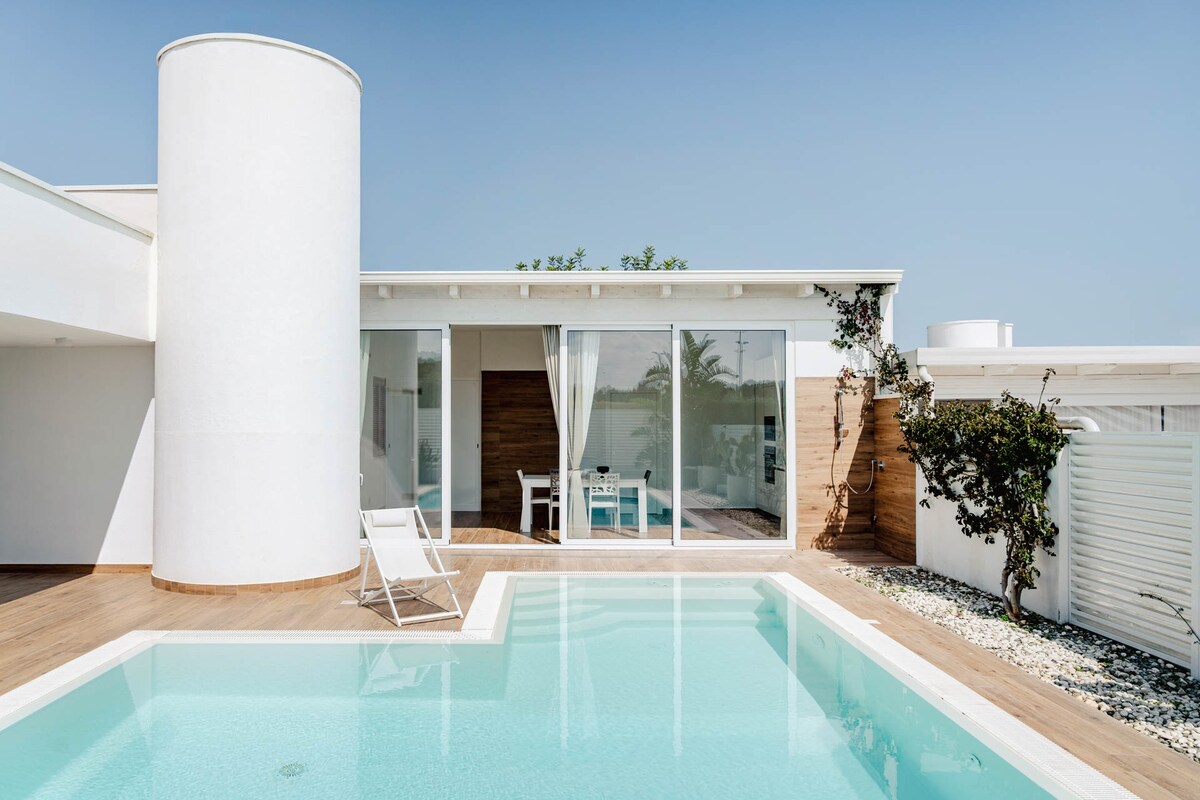 An outdoor view of a modern villa is presented, featuring a rectangular infinity pool and expansive glass doors that lead into the interior. A white seating area is visible through the glass, framed by lush greenery and light-colored wooden accents.