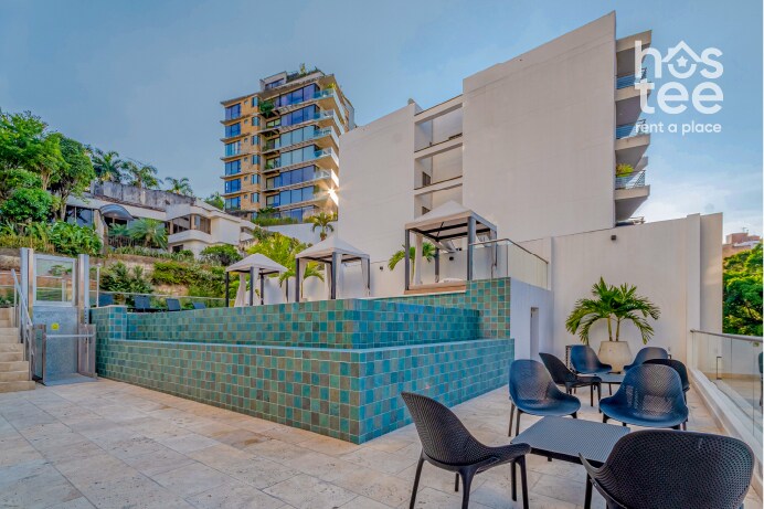 The outdoor area features a landscaped pool with teal tiles, surrounded by several shaded seating areas. Modern lounge chairs are arranged around the pool, and tall greenery provides a serene backdrop. The bright sky enhances the inviting atmosphere of this shared space.