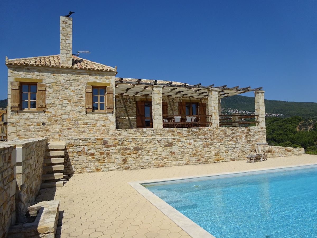 A traditional stone cottage is featured, set against a clear blue sky. The spacious terrace is lined with wooden furniture, providing a view of the pool. Smooth stone steps lead down to the pool area, enhancing the seamless flow between outdoor living and relaxation.