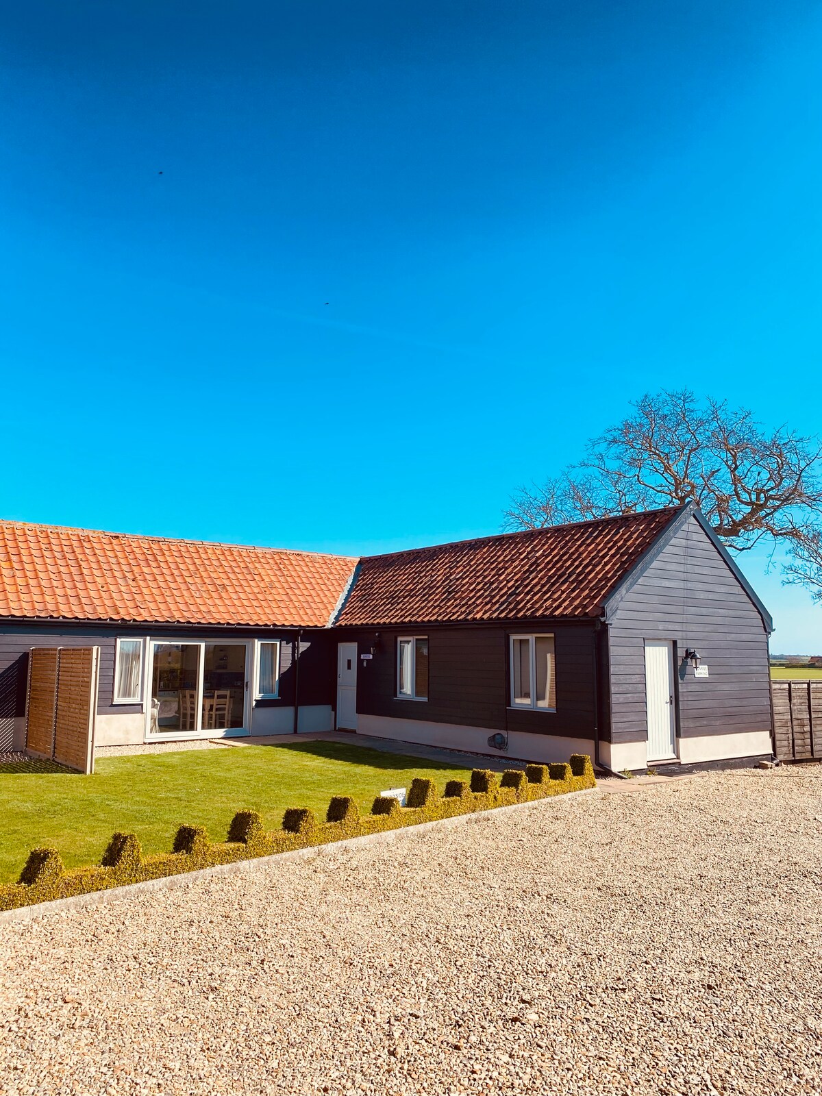 The exterior of the barn conversion is displayed, showcasing a black and grey façade with a red-tiled roof. A well-maintained lawn is visible in the foreground, bordered by small hedges. Clear blue skies create a bright, inviting backdrop for the property.