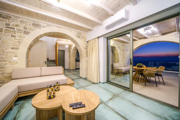 Villa 2: Open-plan living room with modern furnishings, soft tones, and large windows that invite natural light and stunning sea views.