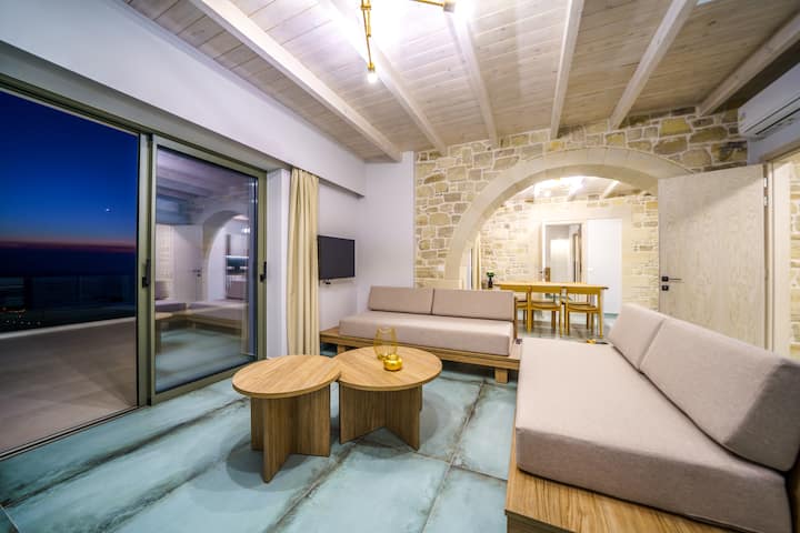 Villa 1: Open-plan living room with modern furnishings, soft tones, and large windows that invite natural light and stunning sea views.